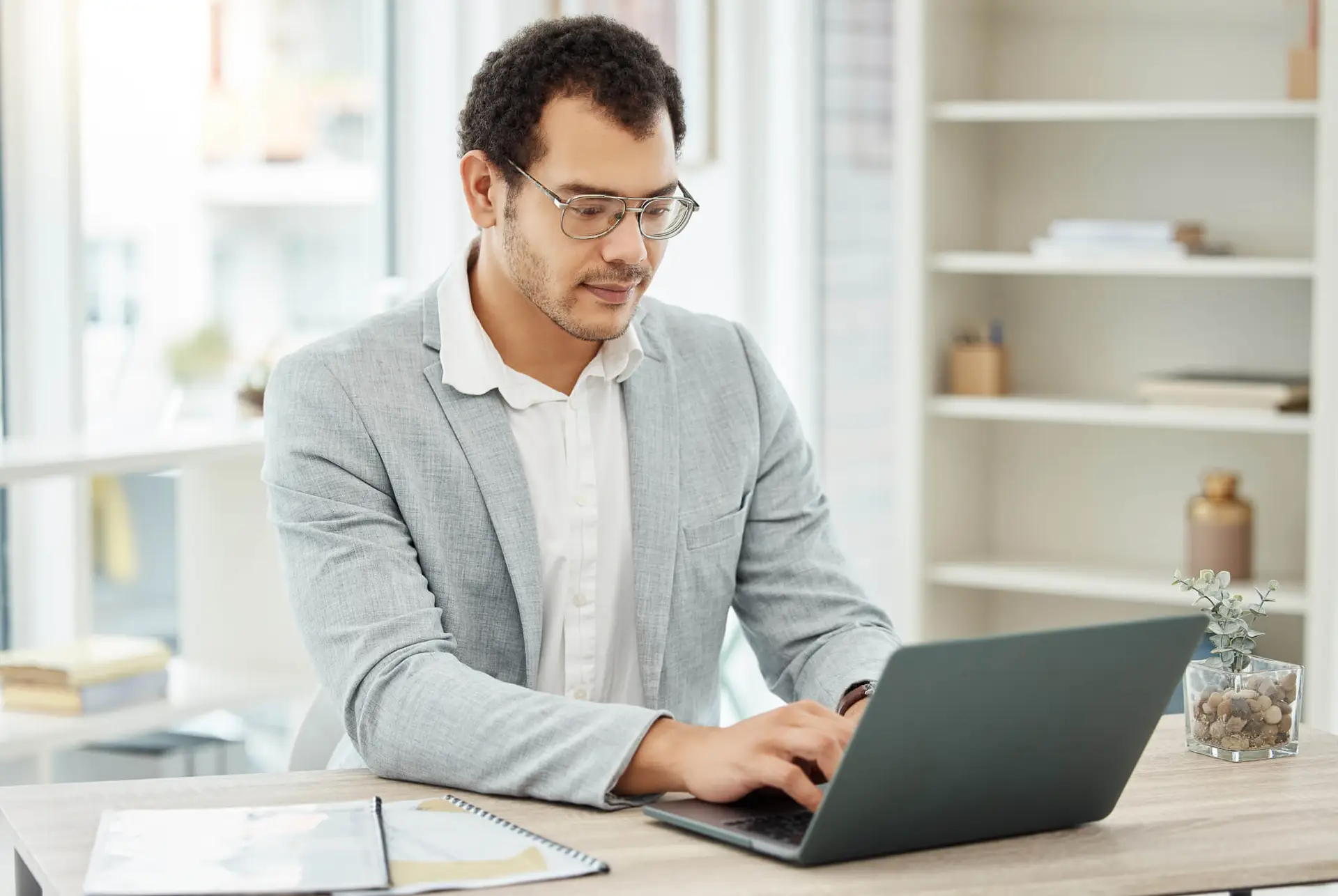 Young professional man in glasses and gray blazer working on a laptop in a bright modern office