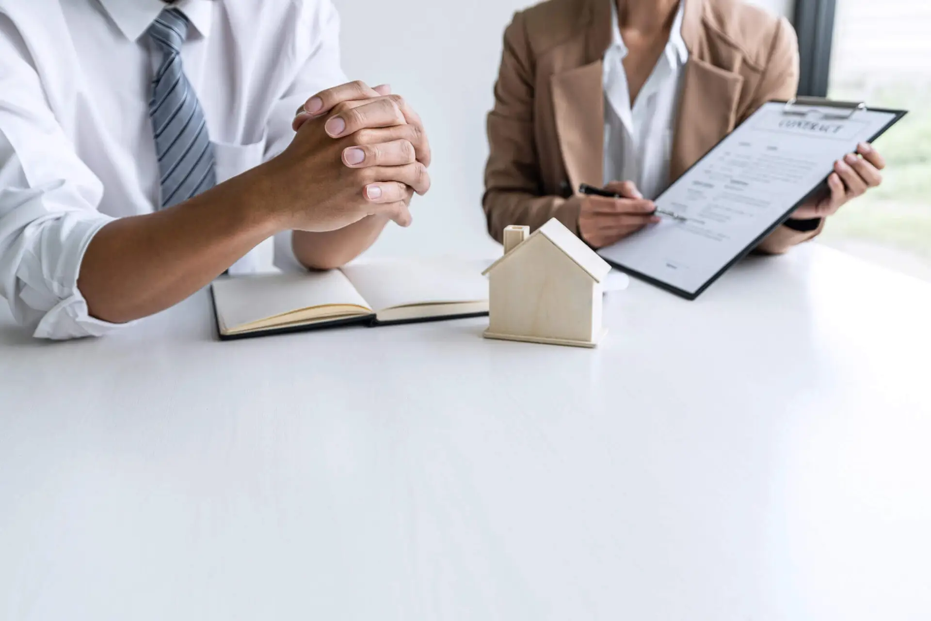 Sebastian Skibinski Two people sit at a desk with a notebook, a small wooden house model, and a clipboard holding a contract. Mortgage Agent