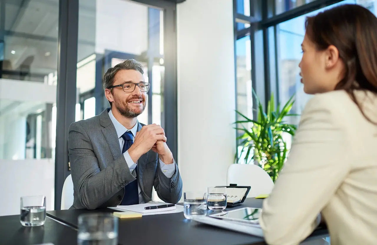Suited man with glasses meets woman at table in modern windowed office.