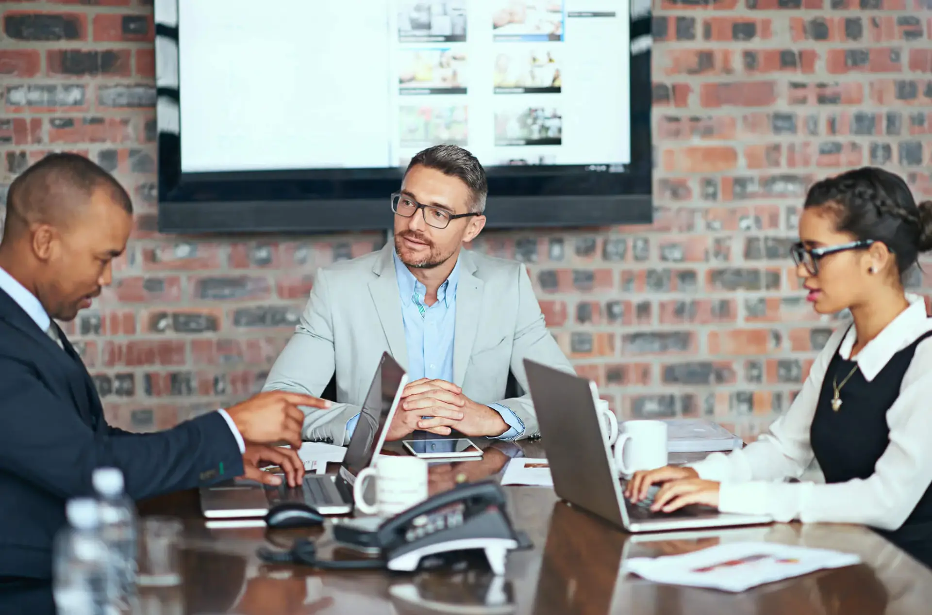 Sebastian Skibinski Three people in business attire sit at a conference table with laptops, having a meeting; a large screen and exposed brick wall are visible in the background. Mortgage Agent