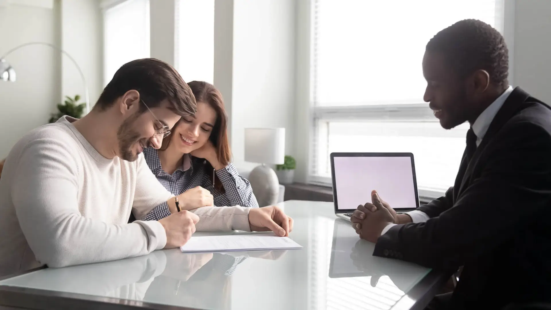 Man signs document beside woman across from suited professional in bright office.