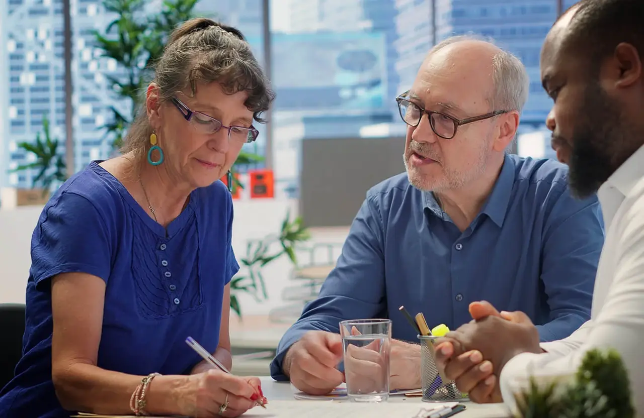 Three adults at office table discussing paperwork; one woman writes while two men converse.
