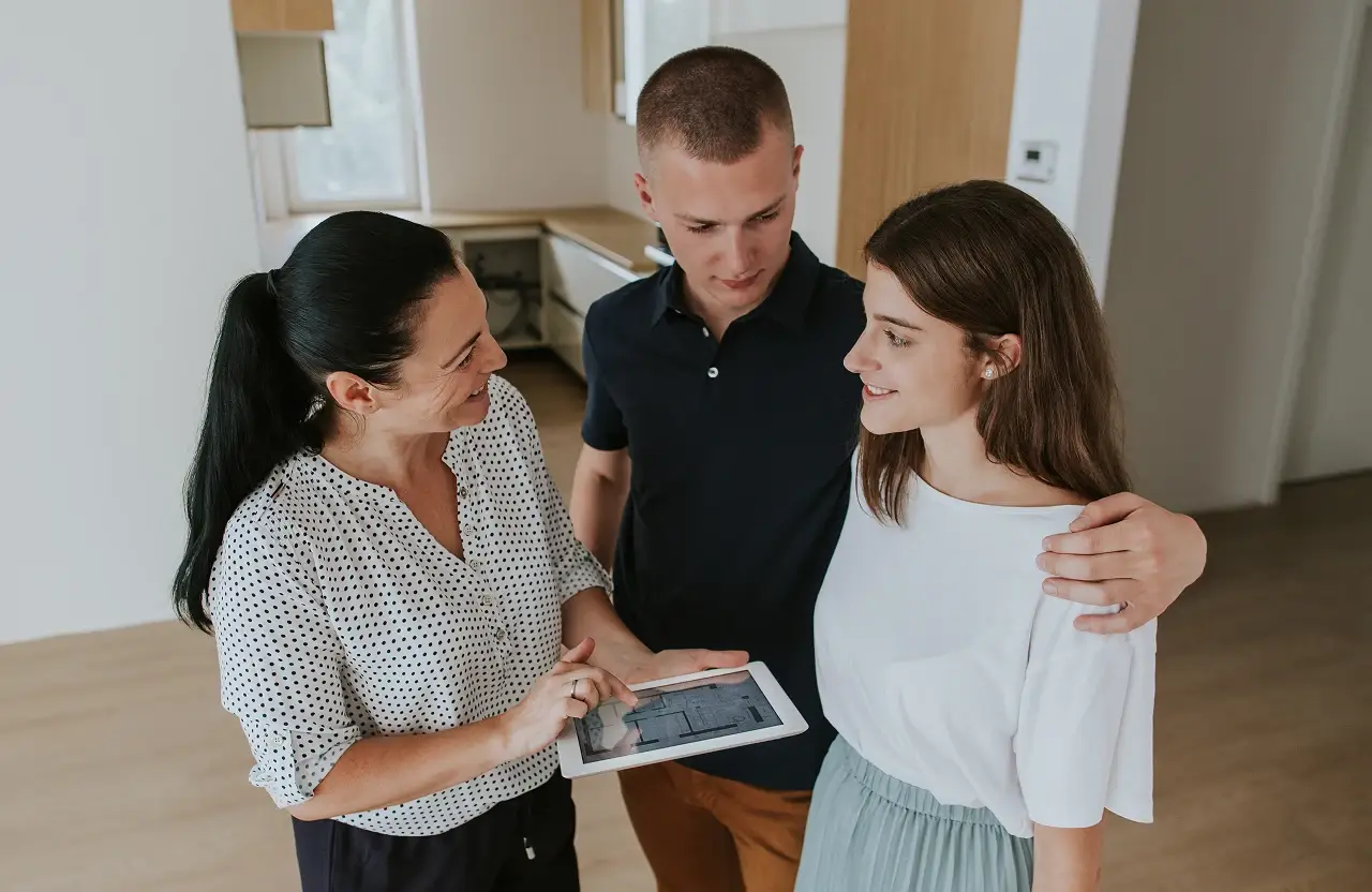 Three people in a modern unfurnished room reviewing a tablet together attentively.