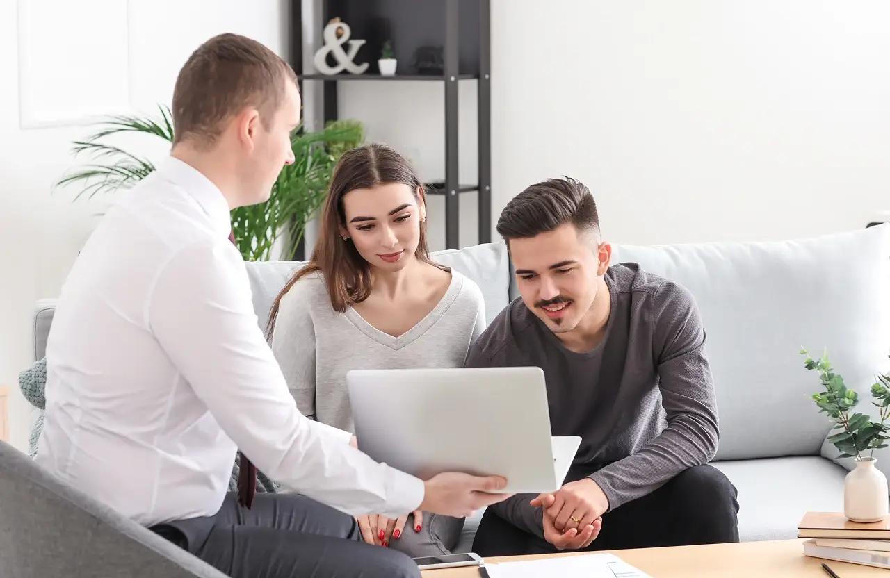 Three people review laptop together on couch in bright modern living room with paperwork.