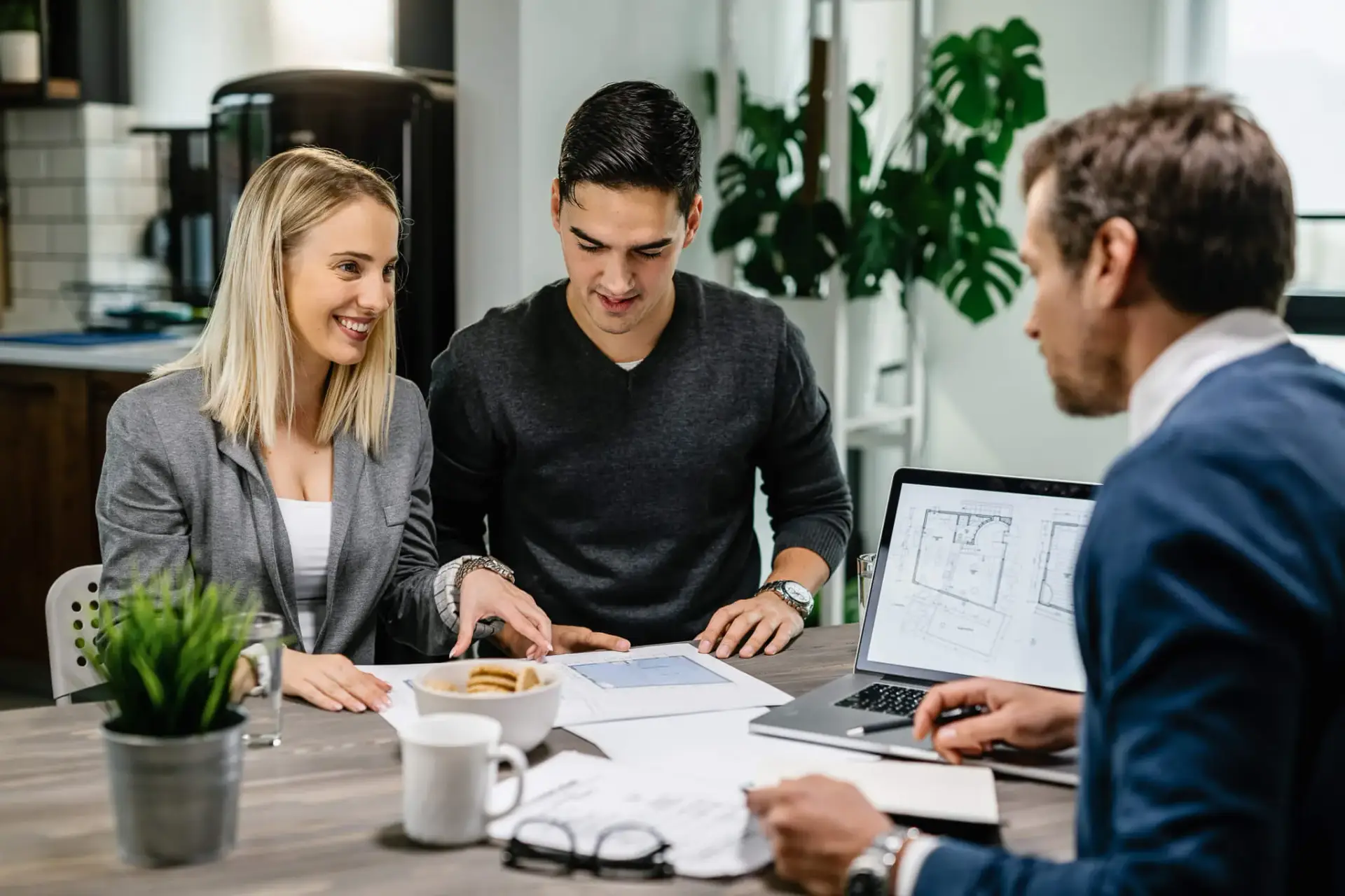Three people discuss documents with architectural plans on laptop over coffee in modern office.